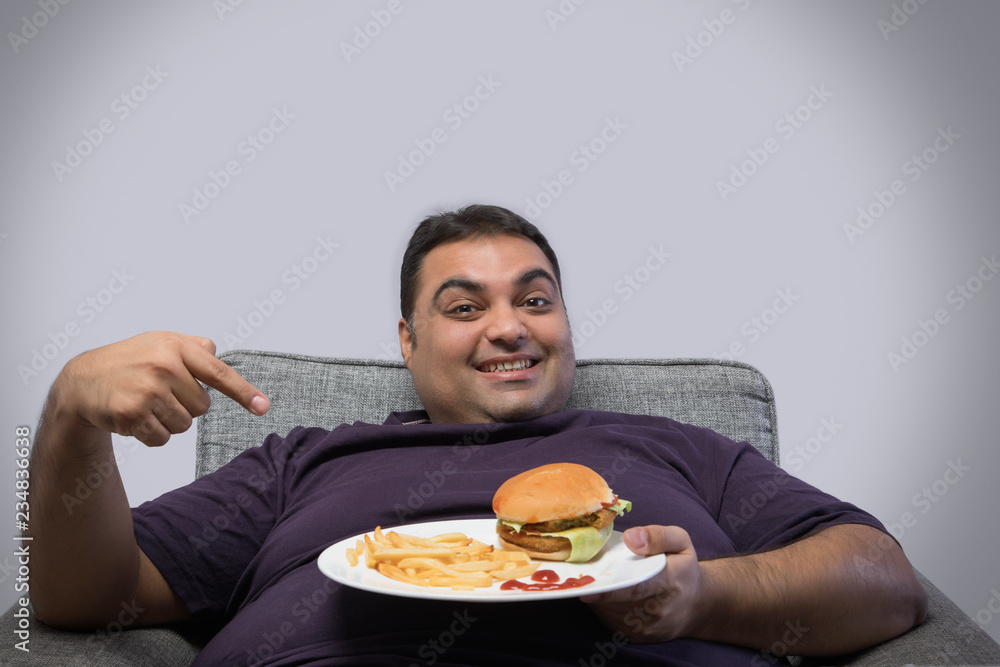 Smiling obese man sitting on chair holding a plate with burger and french fries pointing at it happily Smiling obese man sitting on chair holding a plate with burger and french fries pointing at it happily