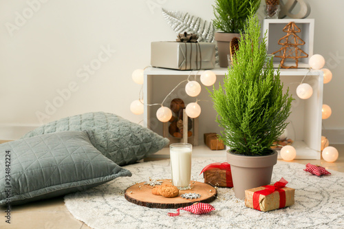 Glass of milk with tasty Christmas cookies and thuya tree on floor in room photo