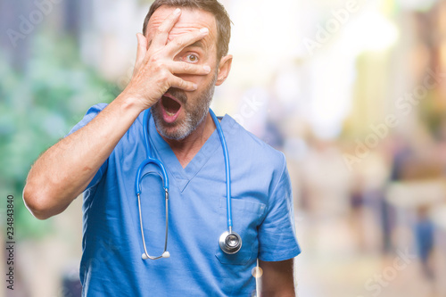 Middle age hoary senior doctor man wearing medical uniform over isolated background peeking in shock covering face and eyes with hand, looking through fingers with embarrassed expression.