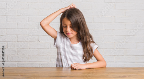 Young hispanic kid sitting on the table at home confuse and wonder about question. Uncertain with doubt, thinking with hand on head. Pensive concept.