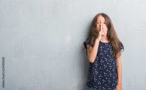 Young hispanic kid over grunge grey wall asking to be quiet with finger on lips. Silence and secret concept.