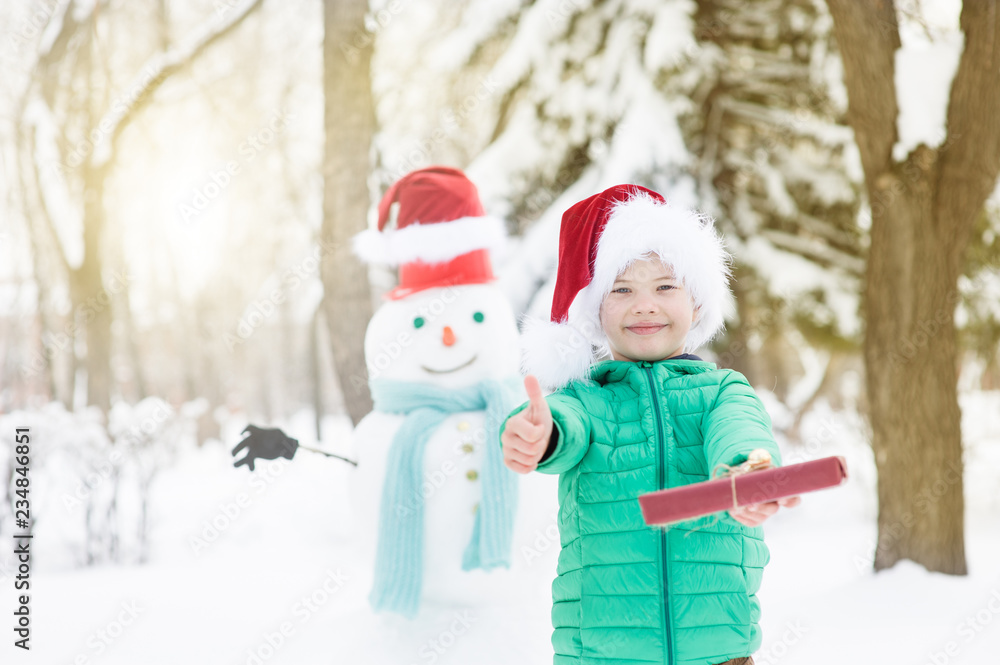 Smiling boy in red christmas hat with snowman on background holds gift box and showing thumbs up