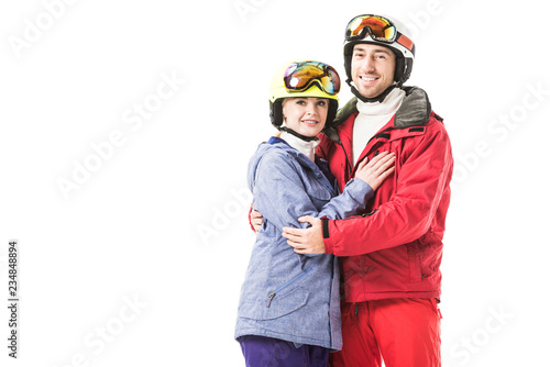 Couple in ski suits, goggles and helmets hugging, smiling and looking at camera isolated on white