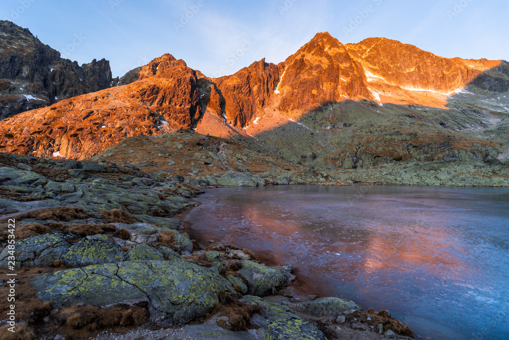 Sunrise photograph of a red-colored mountain massif towering over the frozen surface of a lake