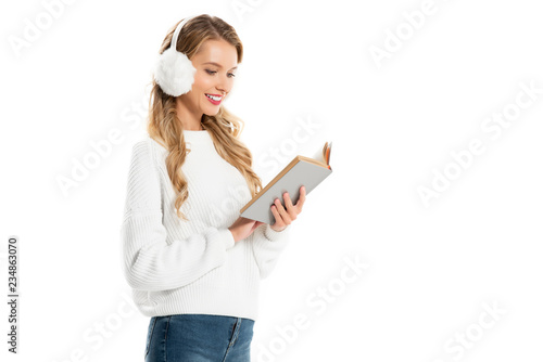 cheerful attractive woman in white earmuffs reading book isolated on white