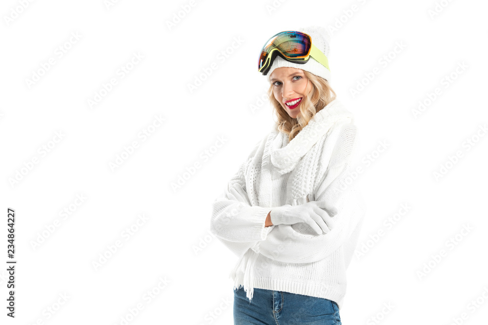 Beautiful young woman in warm clothes and ski goggles with arms crossed smiling and looking at camera isolated on white
