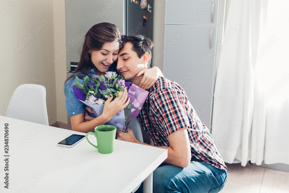 Young man gifting bouquet of flowers to his girlfriend in kitchen. Happy couple hugging. Romantic surprise Young man gifting bouquet of flowers to his girlfriend in kitchen. Happy couple hugging. Romantic surprise