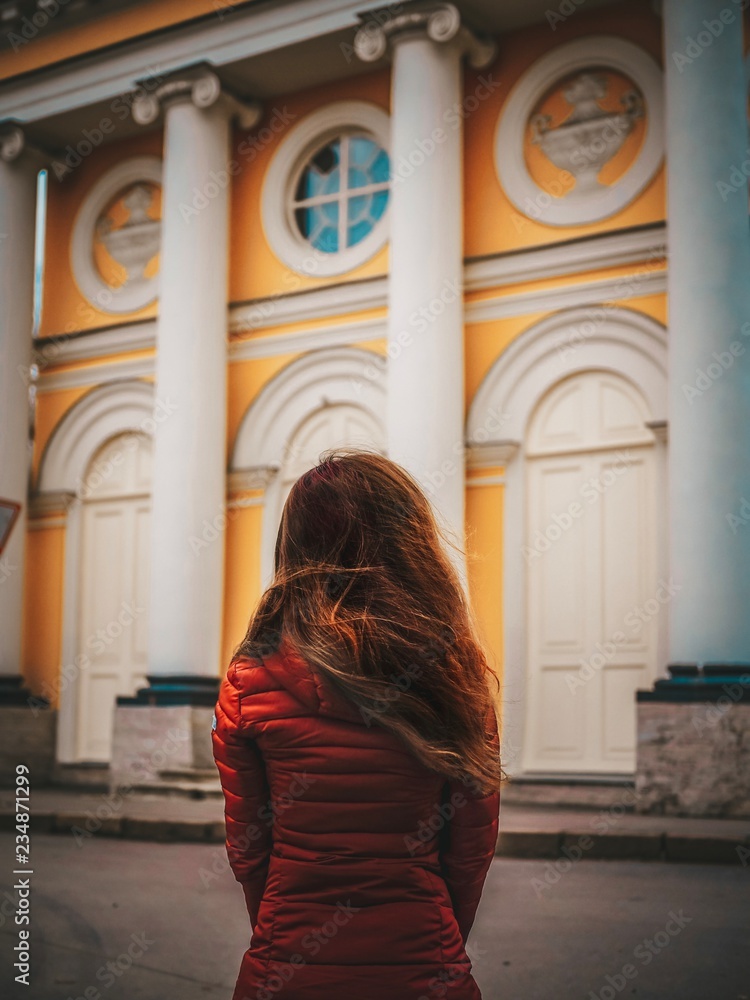 Brunette girl with long hair, in a jacket stands back to the camera and looks at the beautiful old Church with yellow stucco facades