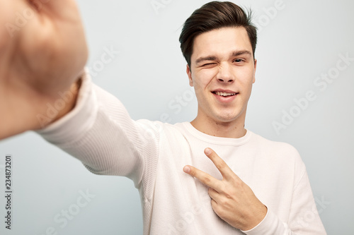 Funny dark-haired guy dressed in a white long sleeve t-shirt shows a v sign taking selfie on a white background