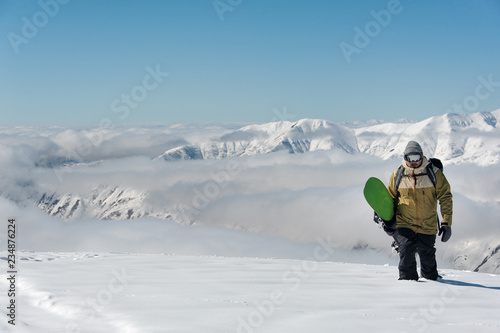 Manful snowboarder walking with the snowboard on the background of mountains