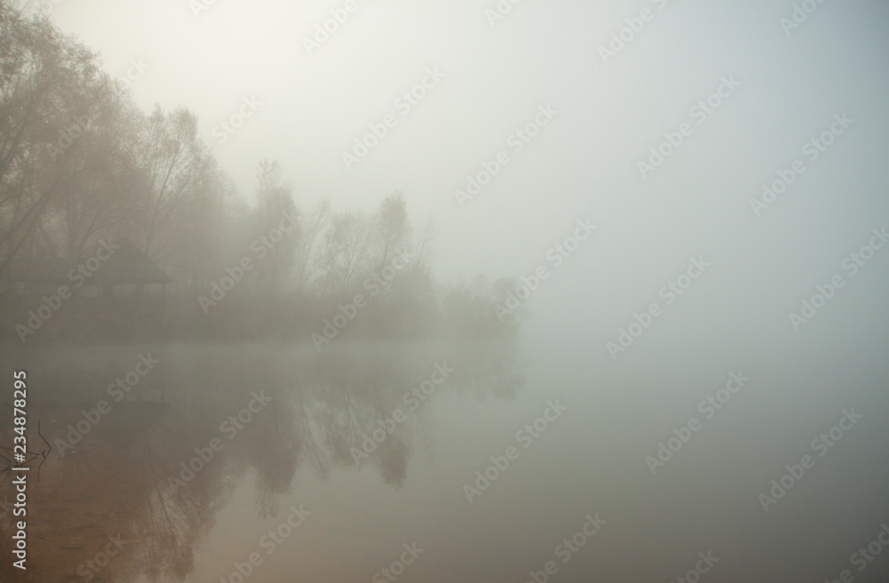 Fototapeta premium Misty morning on the lake. Gazebo or hunting lodge in the forest by the lake. Trees and grass near water. Calm autumn landscape.