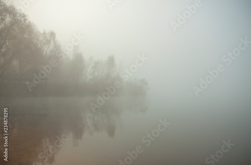 Misty morning on the lake. Gazebo or hunting lodge in the forest by the lake. Trees and grass near water. Calm autumn landscape.