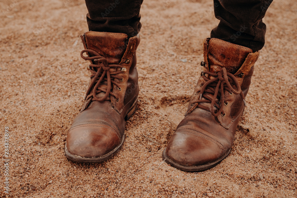 Image of unrecognizable man stands on sandy ground in brown leather old shaggy shoes with laces. Mens footwear. Outdoor shot