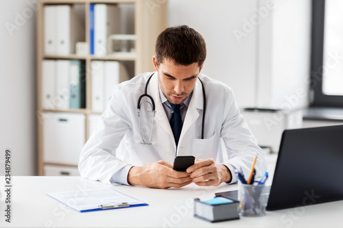 healthcare, medicine and technology concept - male doctor with smartphone at medical office in hospital