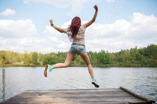 Photo from back of young jumping woman on wooden bridge by river