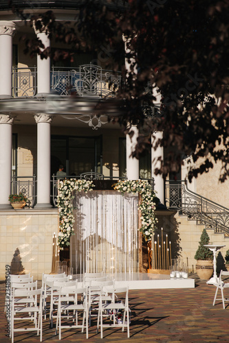 Weddind decoration on open air. Floral decor of a beautiful white and gold arch. Beautiful beckground view of restaurant and trees. Stool for ceremony