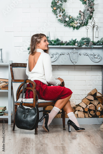 gorgeous young female in a red skirt and white pullover sitting in retro armchair on a christmas decorated wall background. Erotic legs in white socks and black varnish high heells photo