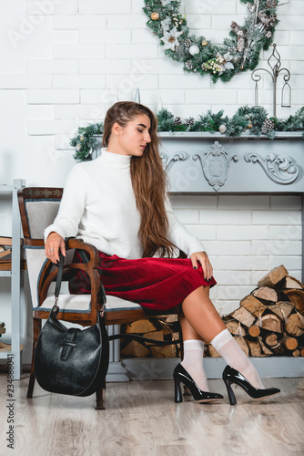 gorgeous young female in a red skirt and white pullover sitting in retro armchair on a christmas decorated wall background. Erotic legs in white socks and black varnish high heells photo