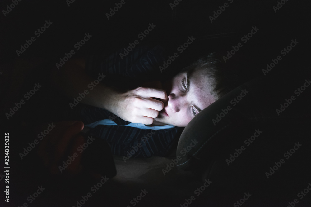 Curious teenager browsing the internet on his mobile phone as he is lying on a couch in the dark. Curious teenager browsing the internet on his mobile phone as he is lying on a couch in the dark.