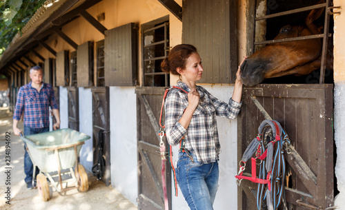 Mature positive female farmer standing near horse at stable
