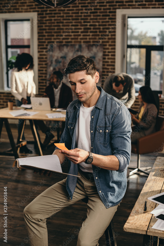 young man holding journal and looking at note in modern loft office with colleagues working on background