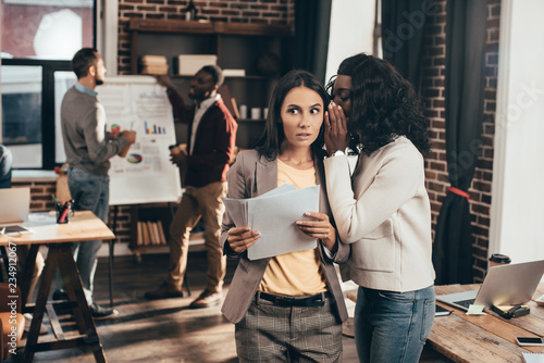 multiethnic couple of business women whispering in loft office with colleagues on background