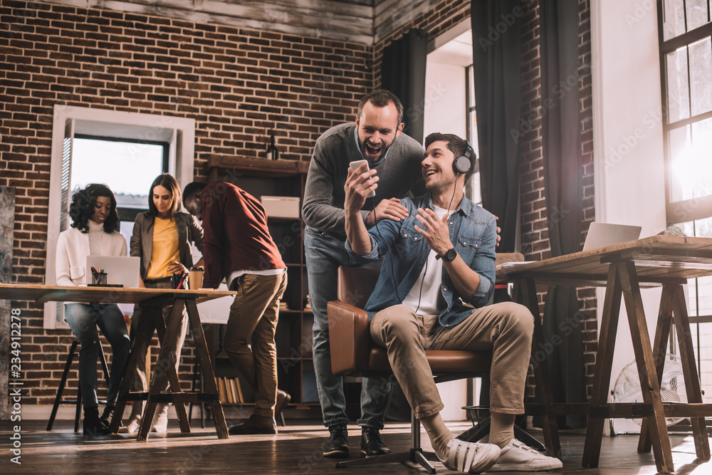 Fototapeta premium two smiling men using smartphone with multiethnic group of casual businesspeople in modern loft office behind