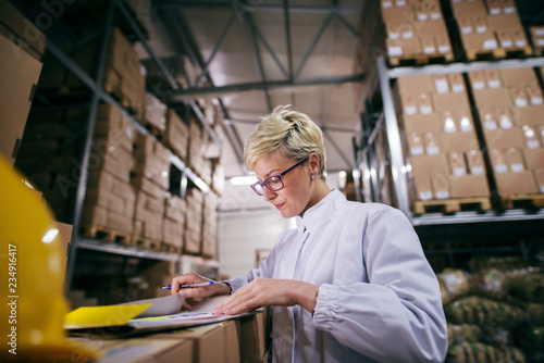 Close up of woman filling paperwork while standing in warehouse. In background shelves with boxes.