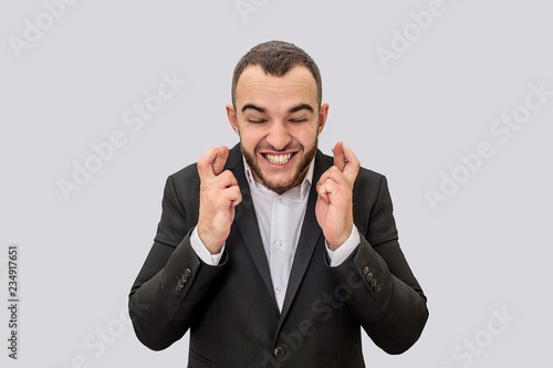 Young businessman stands and keeps fingers crossed. He shrinks and smiles. Isolated on white background.