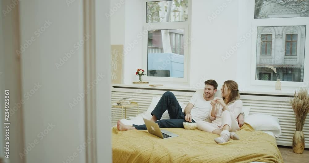 Weekend time for a young couple in pajamas spending all day in bed eating popcorn and watching movie on the notebook. 4k