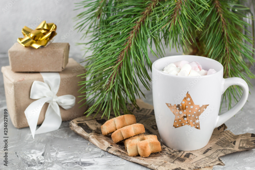 gift boxes,mug with drink decorated with marshmallow and star shape cookies  near evergreen christmas tree branches gray concrete background
