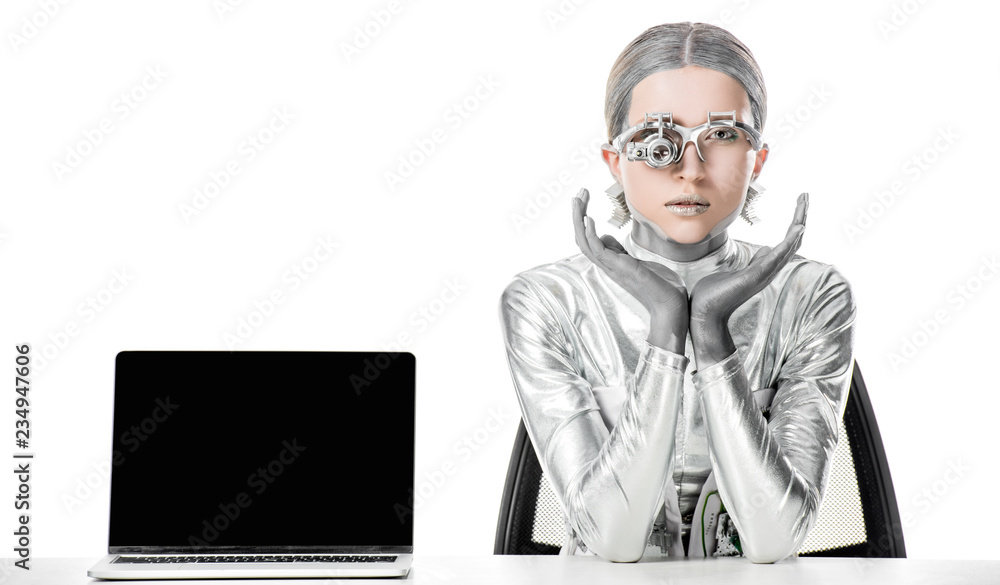 silver robot sitting at table near laptop with blank screen isolated on white, future technology concept silver robot sitting at table near laptop with blank screen isolated on white, future technology concept