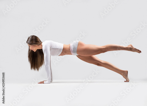 Beautiful brunette fitness model wearing tight, short gray fitness and a white long sleeve sports top does bridge with one leg raised a in studio on a light gray background