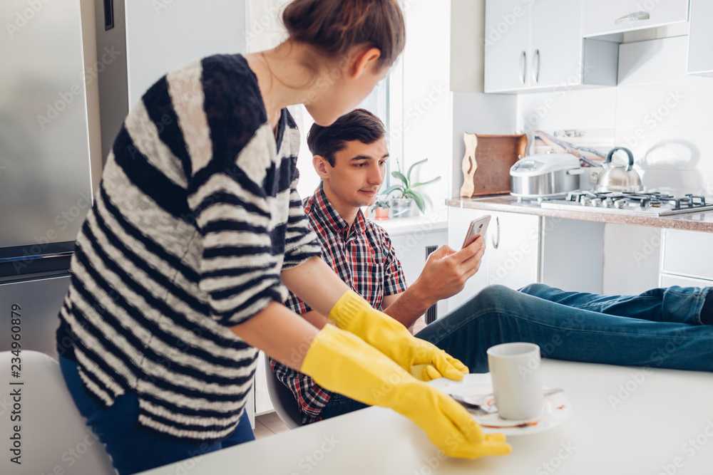 Lazy man looking at phone while his angry girlfriend cleaning his dishes in kitchen and talking to him. Family arguement Lazy man looking at phone while his angry girlfriend cleaning his dishes in kitchen and talking to him. Family arguement