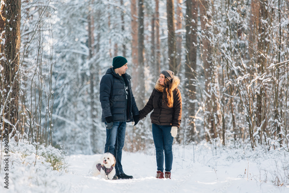 Cute young couple having fun in winter forest with their pretty little white dog. Man and woman in sweater with a snowflake. Christmas and winter holidays
