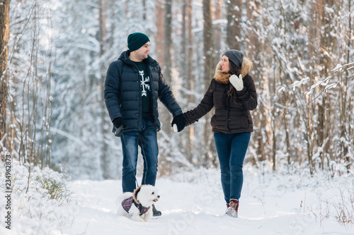 Cute young couple having fun in winter forest with their pretty little white dog. Man and woman in sweater with a snowflake. Christmas and winter holidays
