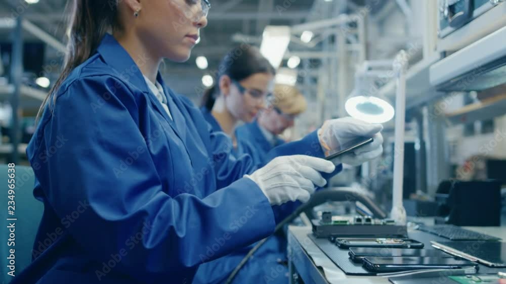 Female Electronics Factory Worker in Blue Work Coat and Protective Glasses Takes a Smartphone Screen and Performs a Quality Check. High Tech Factory Facility with Multiple Employees. Female Electronics Factory Worker in Blue Work Coat and Protective Glasses Takes a Smartphone Screen and Performs a Quality Check. High Tech Factory Facility with Multiple Employees.