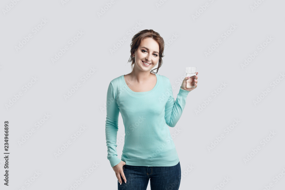 Beautiful girl with brown-haired woman dressed in jeans and mint-colored sweater on a light background holds in her left hand a glass of pure drinking water and smiling. Beautiful girl with brown-haired woman dressed in jeans and mint-colored sweater on a light background holds in her left hand a glass of pure drinking water and smiling.
