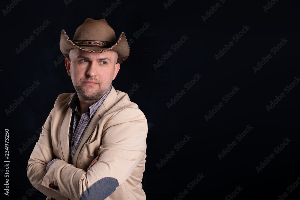 Dramatic portrait of men in beige cowboy's hat on dark background in studio. He stands in a beige jacket with arms crossed. Dramatic portrait of men in beige cowboy's hat on dark background in studio. He stands in a beige jacket with arms crossed.