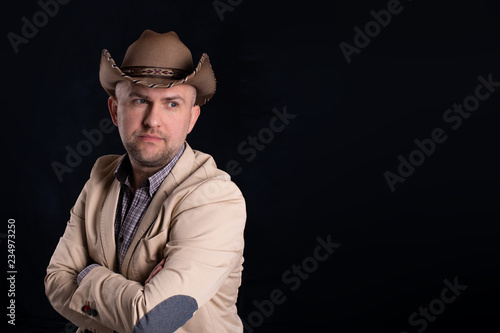 Dramatic portrait of men in  beige cowboy's hat on dark background in studio. He stands in a beige jacket with arms crossed.