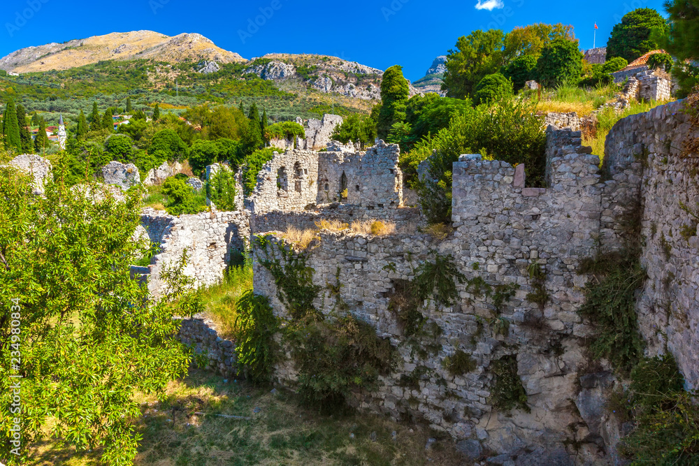 The picturesque historic walls of the destroyed city covered with green plants grass trees on the backdrop of colorful mountains and clear sky. Summer landscape in Fortress Old Bar Town, Montenegro.