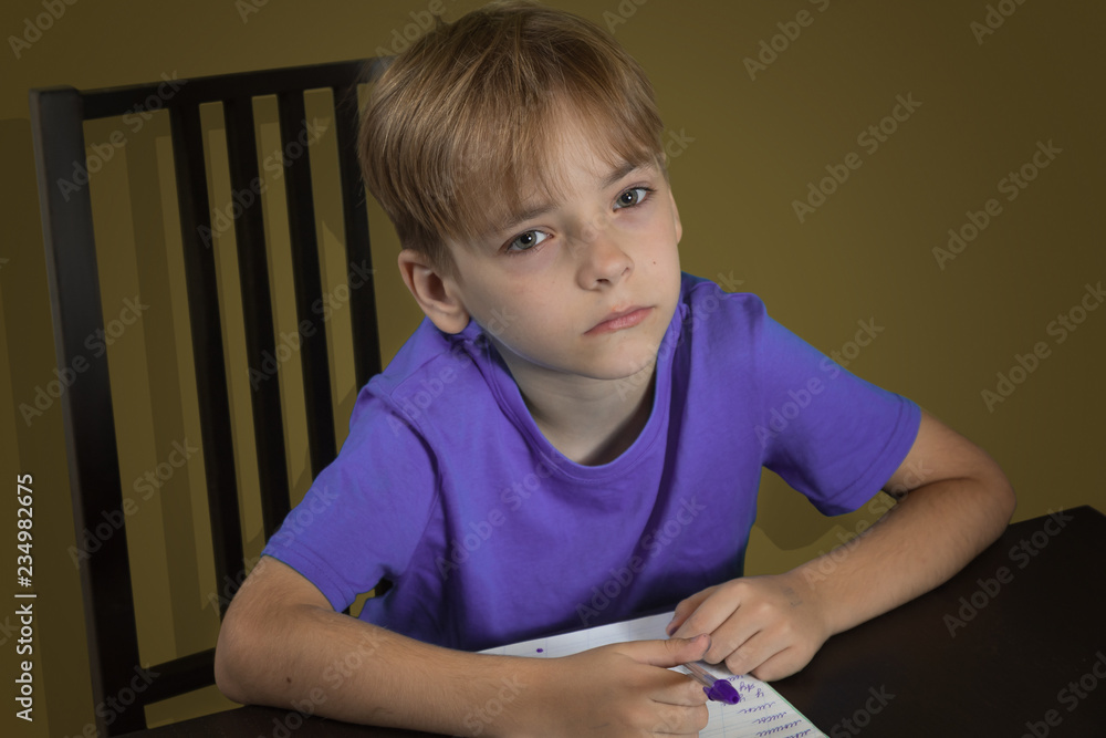 Caucasian boy the pupil of elementary school at a table does homework. The tired and tortured, exhausted child Caucasian boy the pupil of elementary school at a table does homework. The tired and tortured, exhausted child