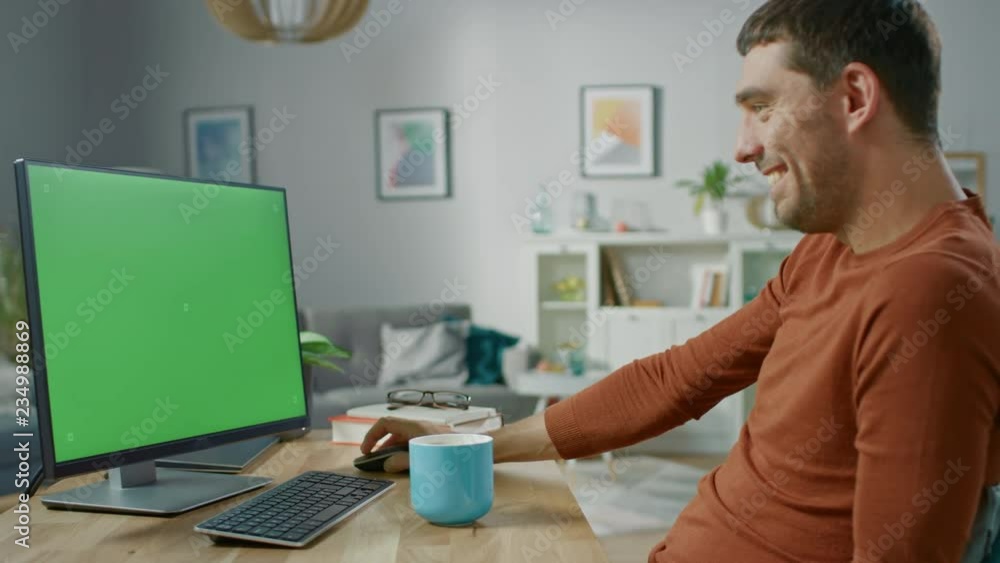 Handsome Smiling Man Sitting at His Desk at Home Uses Personal Computer with Mock-up Green Screen. He Lives in Cozy Aparment with Modern Living Room Interior. Handsome Smiling Man Sitting at His Desk at Home Uses Personal Computer with Mock-up Green Screen. He Lives in Cozy Aparment with Modern Living Room Interior.
