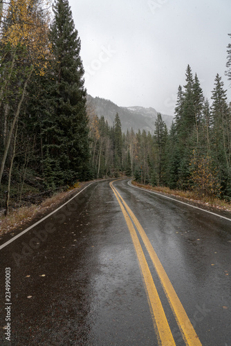 A paved mountain road cuts through evergreen forest and yellow and orange aspens of autumn as the moody first snow descends on the mountains