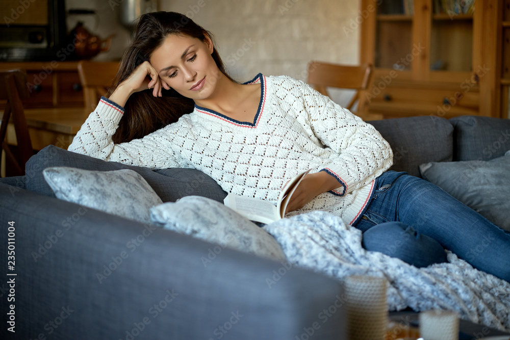 Obraz premium beautiful young brunette woman reading a book on a gray sofa.
