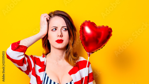 Portrait of beautiful young surprised redhead white woman in red striped shirt with heart shape toy on yellow background