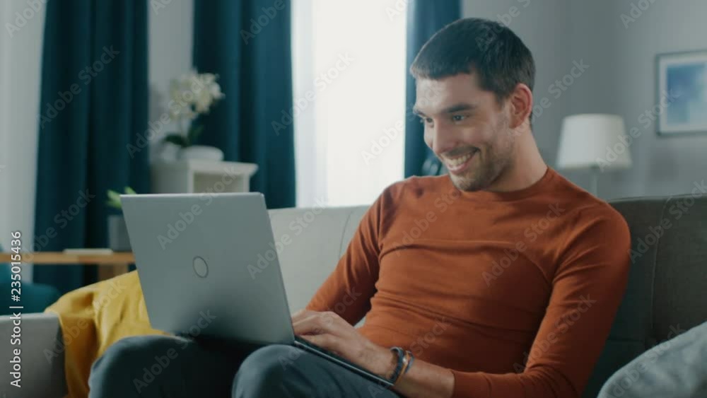 Handsome Young Man Holding Laptop Sits Down on Sofa at Home. Guy Working on Computer from His Cozy Living Room. Handsome Young Man Holding Laptop Sits Down on Sofa at Home. Guy Working on Computer from His Cozy Living Room.
