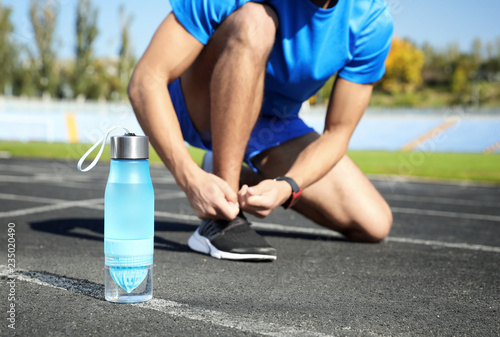 Sporty man tying shoelaces near bottle of water at stadium on sunny day