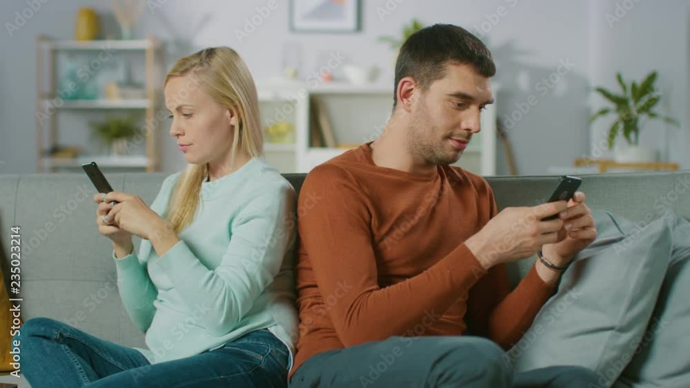 Alienation / Quarrel Concept: At Home on a Sofa Young Couple Sitting Apart, Facing Different Directions and Using Smartphones. Alienation / Quarrel Concept: At Home on a Sofa Young Couple Sitting Apart, Facing Different Directions and Using Smartphones.