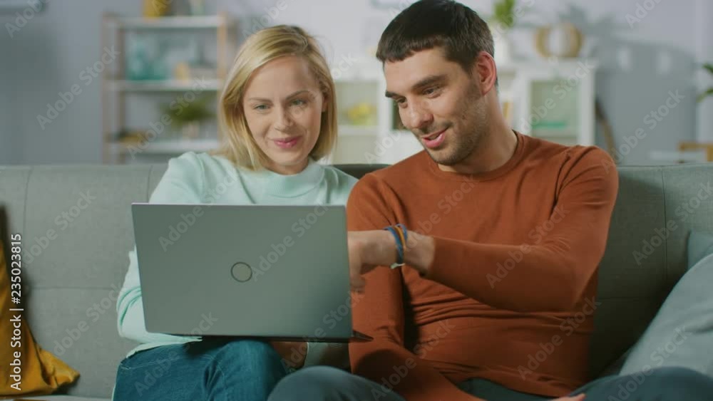 Cute Young Couple Sits on a Sofa at Home, They Look use Laptop Computer and Have Fun. Portrait of Beautiful Happy Young Couple in Love. Cute Young Couple Sits on a Sofa at Home, They Look use Laptop Computer and Have Fun. Portrait of Beautiful Happy Young Couple in Love.
